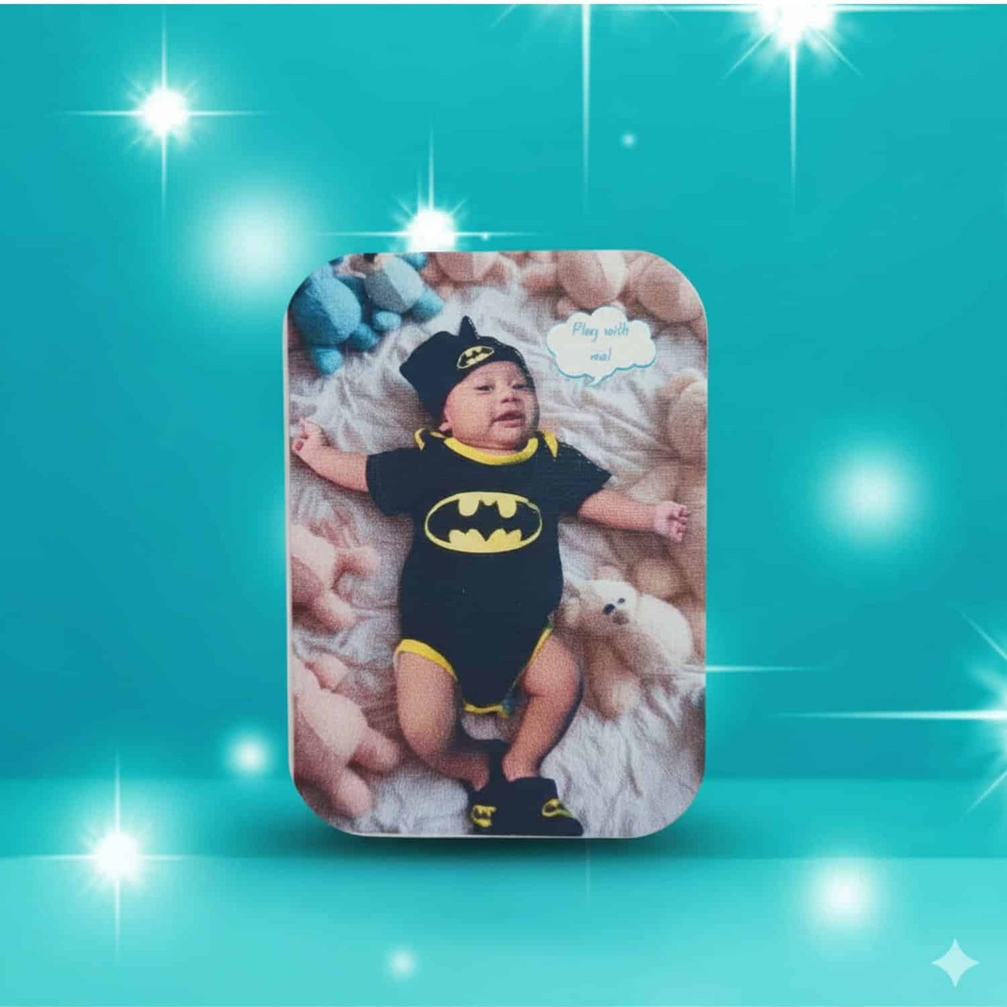 Personalized rounded rectangle magnet featuring a baby dressed in a black and yellow Batman costume and matching hat, lying on a white blanket surrounded by small teddy bears, with the text 'Play with me!' in a speech bubble, on a sparkling teal background.