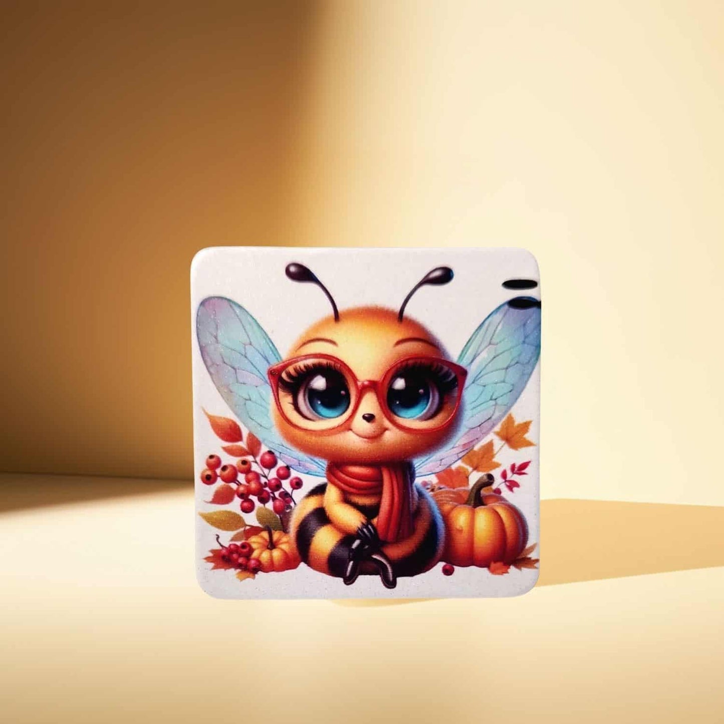 A rounded square magnet, featuring a cute cartoon bee in red glasses, surrounded by autumn leaves and a pumpkin, displayed on a warm, solid light brown background.