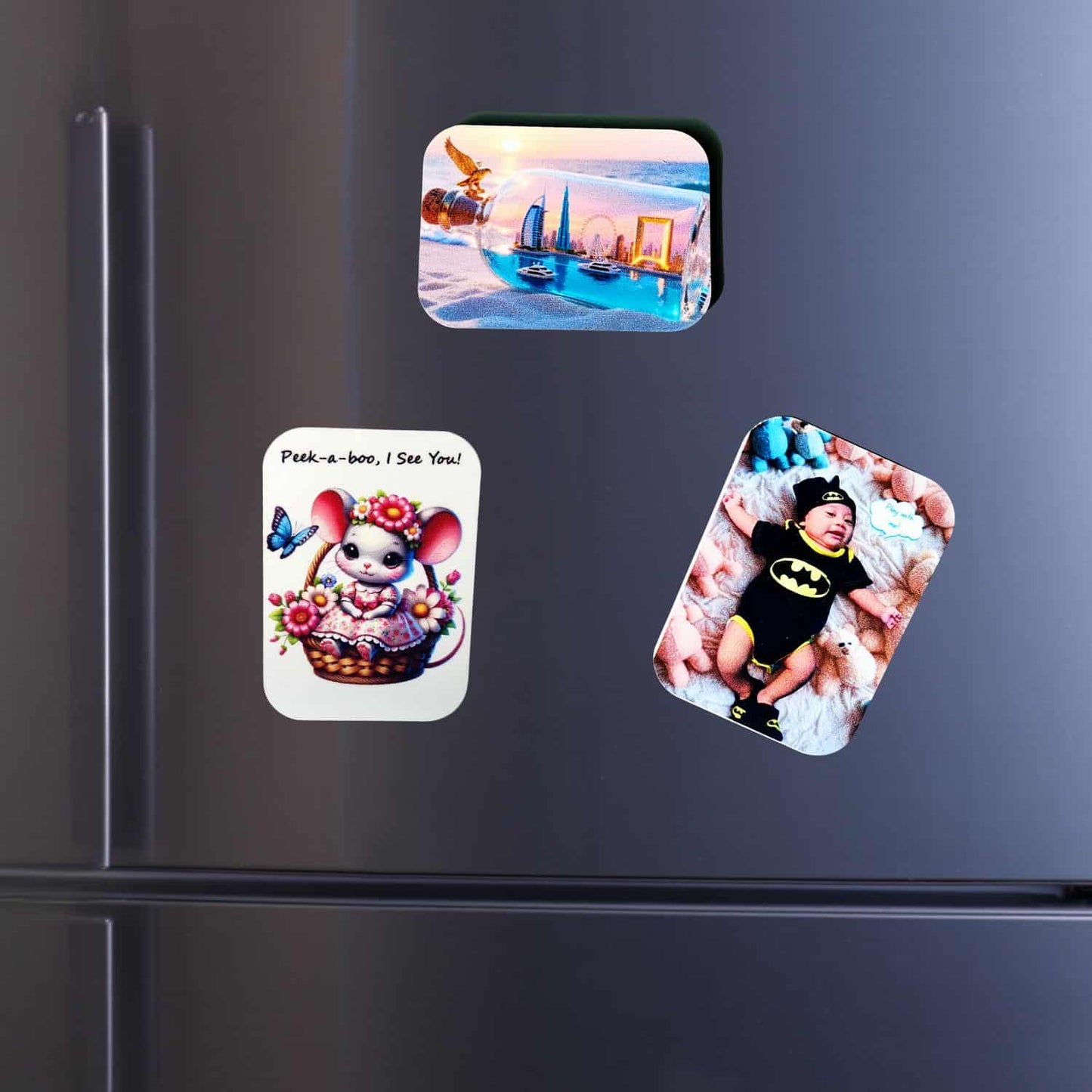 Collection of three custom-printed, rounded rectangle magnets from InkFluenceME on a modern silver fridge. The magnets display a Dubai cityscape, an adorable baby in a Batman onesie, and a cheerful cartoon mouse in a flower basket. This image showcases personalized souvenirs and custom photo magnets.