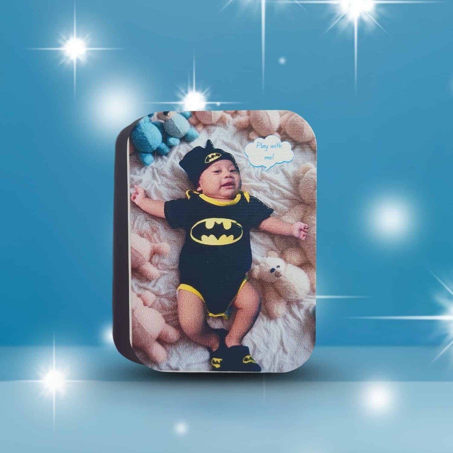InkFluenceME custom rounded rectangle magnet showcasing a heartwarming UV print. The image features a very cute baby dressed in a full black and yellow Batman superhero outfit, complete with the iconic bat-signal on the chest and a pointed hat. The baby is comfortably lying on fluffy, light-colored bedding, surrounded by stuffed toys. This personalized photo magnet is an ideal gift for new parents, baby showers, or as a charming family keepsake.