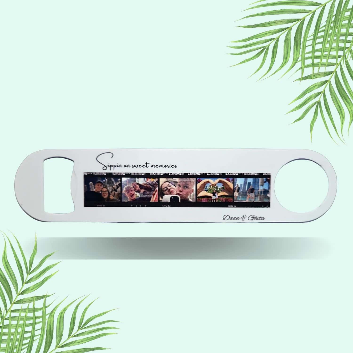 Custom metal bottle opener with a personalized photo film strip and text, "Sippin on sweet memories," laid on a soft teal-green background with subtle palm leaf accents.