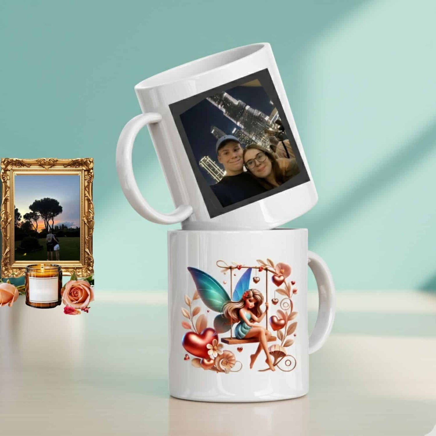 Custom photo mugs stacked, featuring a personalized image of a couple on the top mug and a colorful fairy design on the bottom mug, displayed next to a gold framed picture, a lit candle, and roses on a white table against a soft teal background.